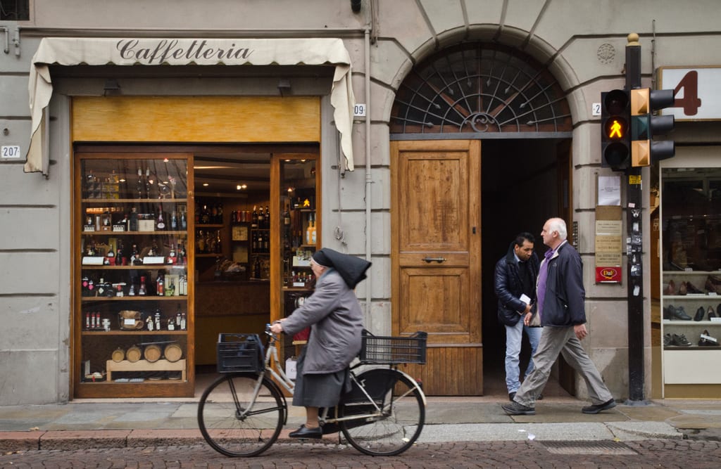 In the city of Modena, a nun wearing a black habit rides by on a bicycle while looking into the open door of a wine shop.