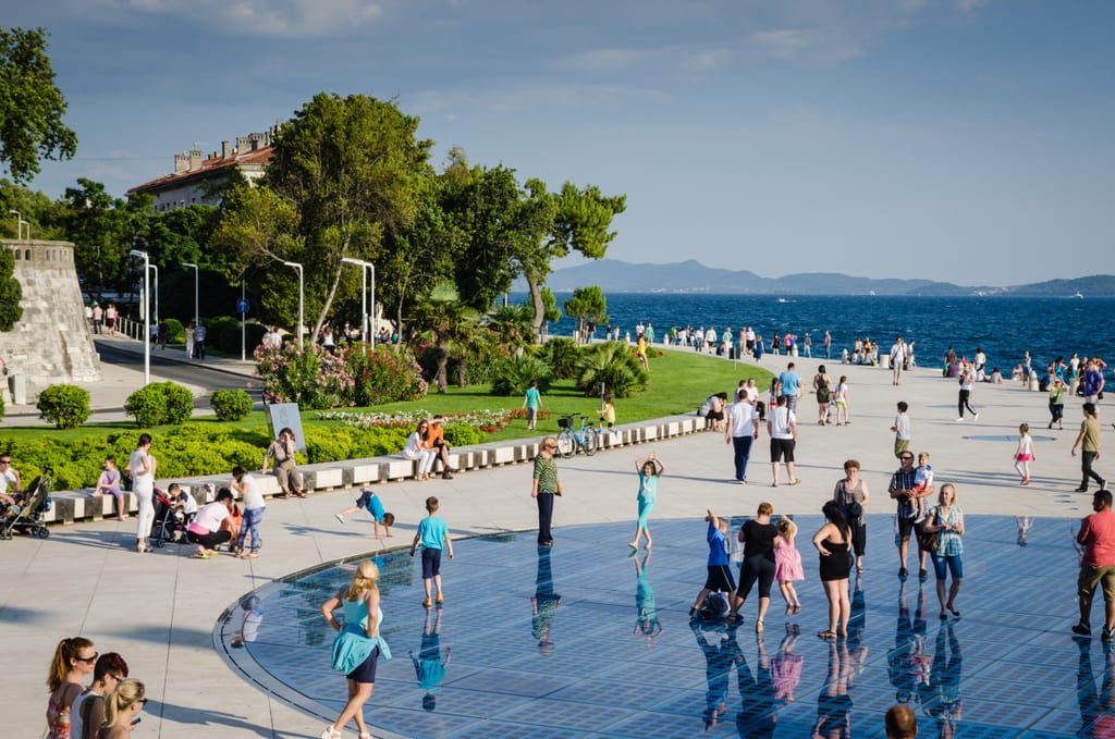 People walking across a blue disk -- the sun salutation -- on the gray boardwalk in Zadar, Croatia, trees on the left side.
