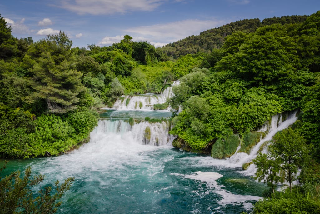 Two giant, multi-tiered waterfalls in Krka National Park overflowing into bright teal water, surrounded by forest.