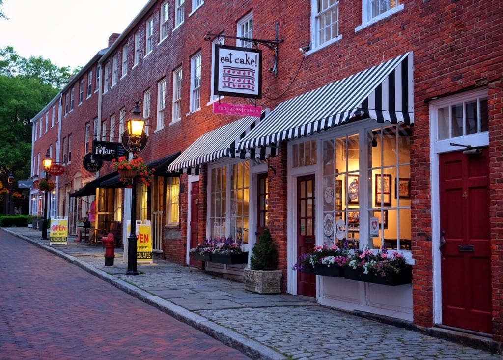A small street in Newburyport, Massachusetts, with brick roads, cobblestone sidewalks, and boutiques including a bakery with an "Eat Cake" sign in a brick building.