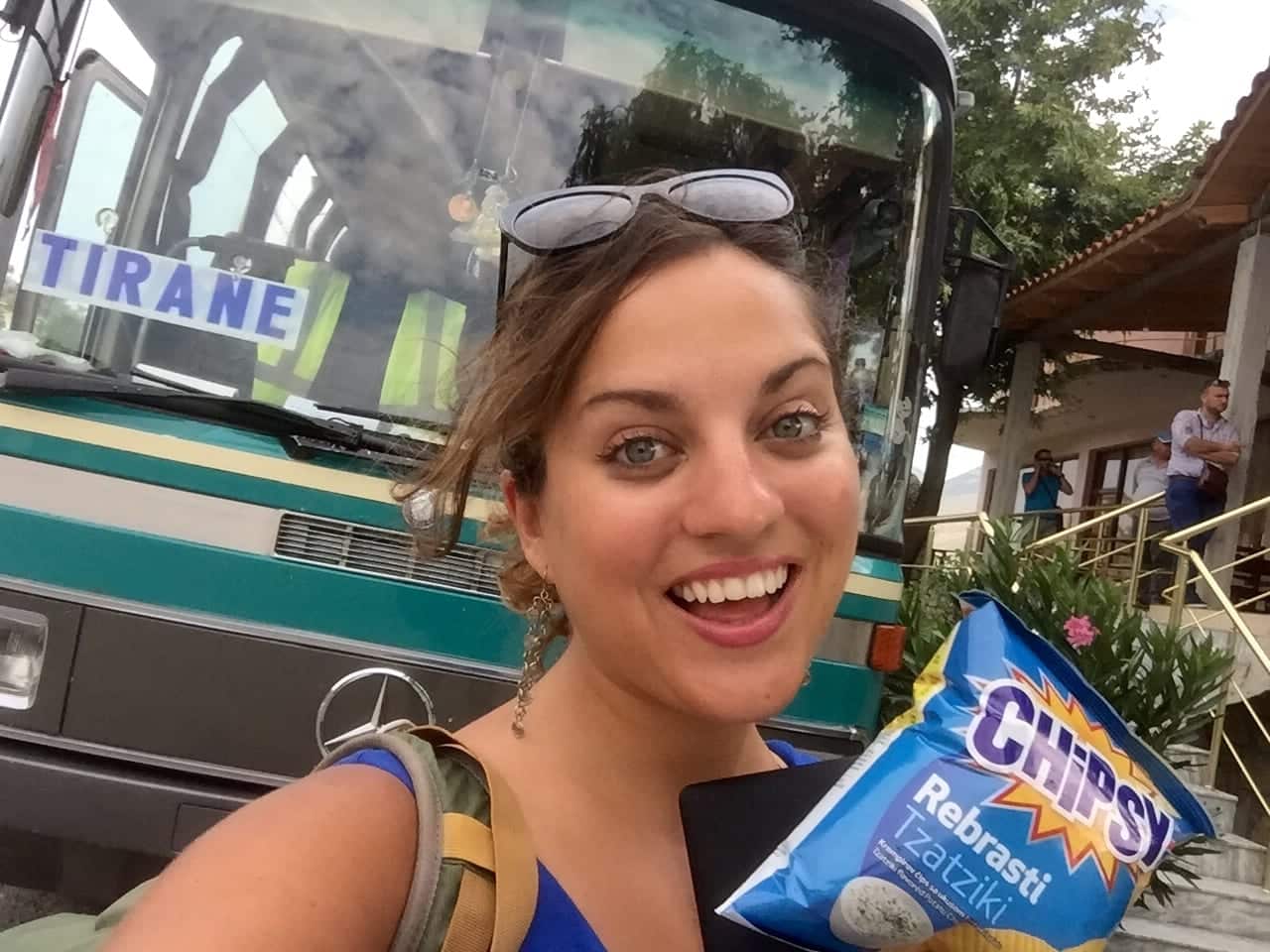 Kate taking a smiling selfie in front of an Albanian bus, holding a pack of Tzatziki chips.