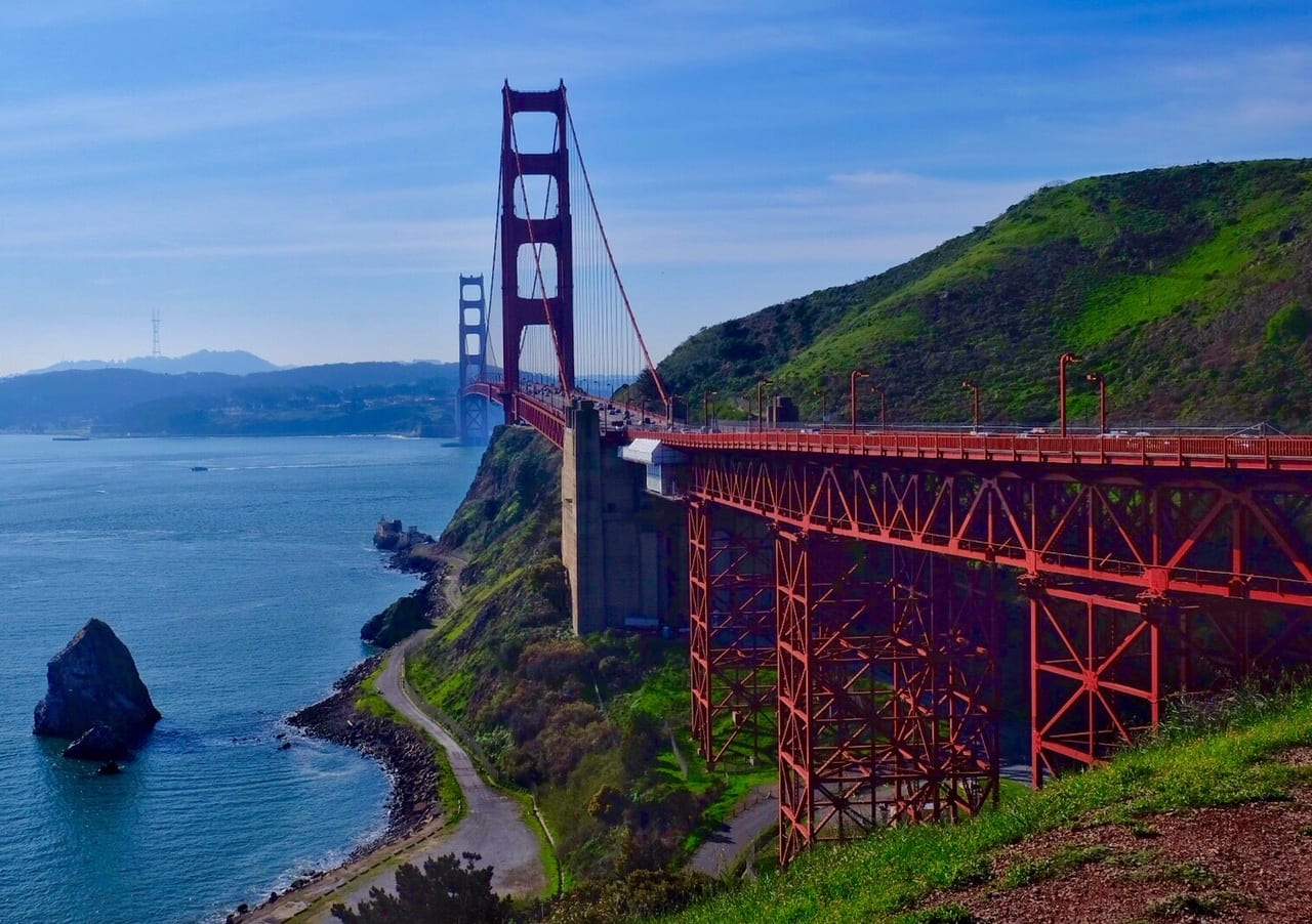 The Golden Gate Bridge rises up from the Sausalito side. It's bright red and extends into the distance, set against green cliffs, above a bright blue ocean, and underneath a streaky blue and white sky. San Francisco is misty in the background.