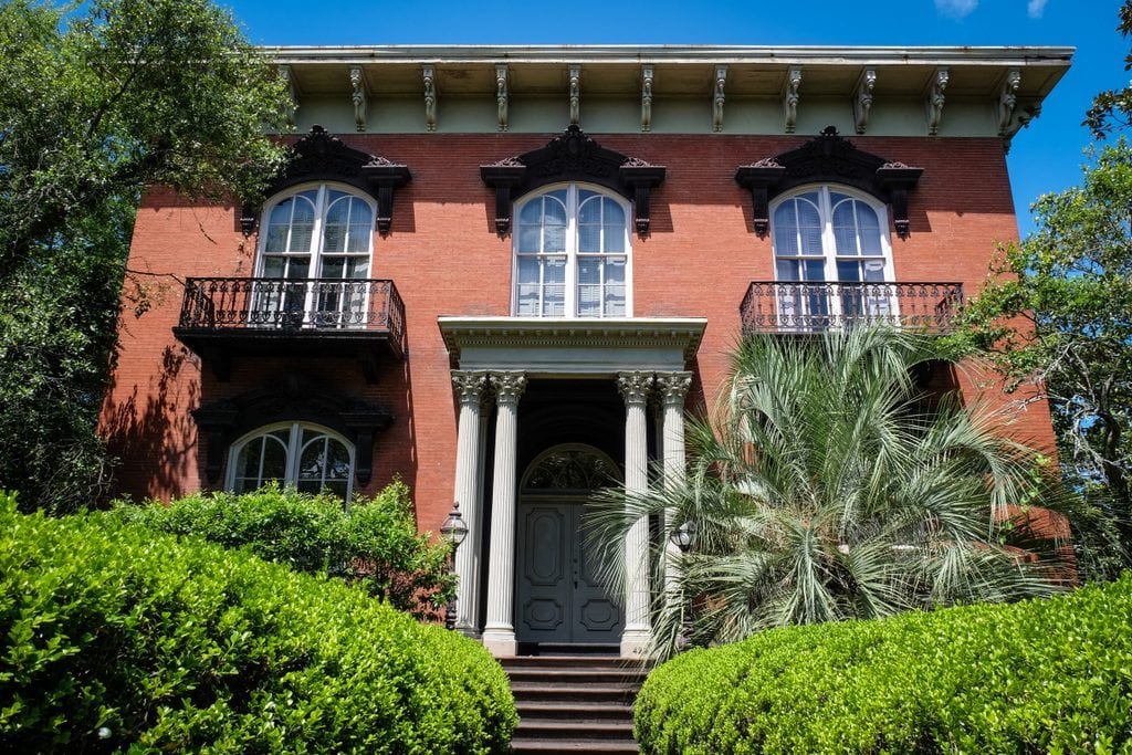 The Mercer House in Savannah: a red brick house with two white columns on each side of the entrance, lots of plants and vegetation in front.