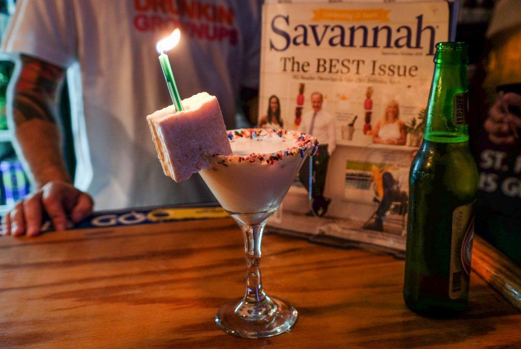 A martini glass with a white liquid inside, rainbow sprinkles on the edge, with a white hostess cake on the rim, a lit green birthday candle sticking out of it.