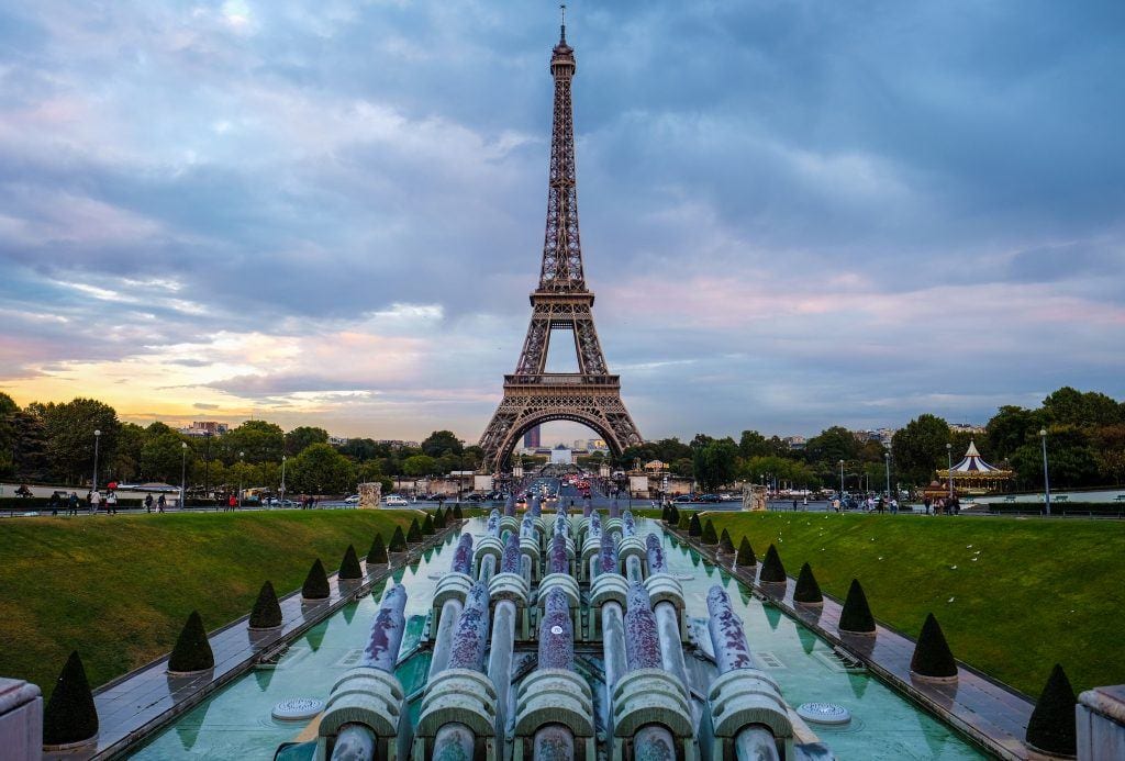 The Eiffel Tower in front of a row of cannons in the Trocadero Gardens, a cloudy sunset behind it.