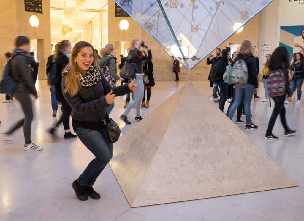 Kate squatting and giving two thumbs up next to the inverted glass pyramid at the Louvre and giggling with glee.