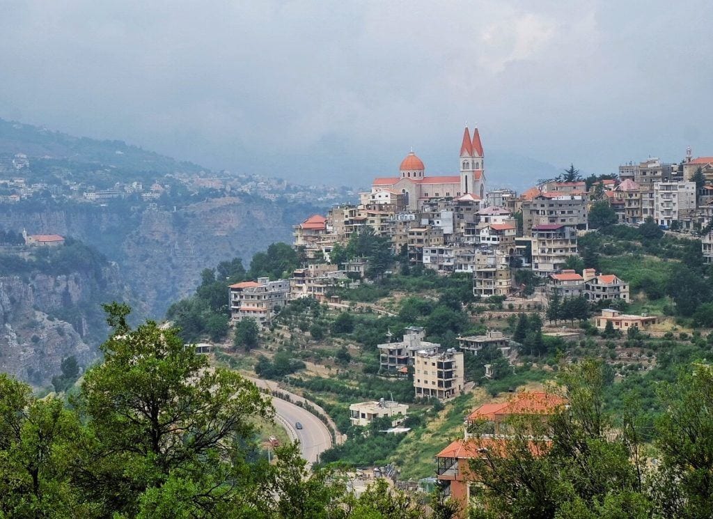 The town of Bcharre, Lebanon, in the distance: you see a small town of white stone and orange roofs perched on a steep hill, greenery poking in between the buildings, a church tower poking out the top. It's a moody day with thick gray clouds.