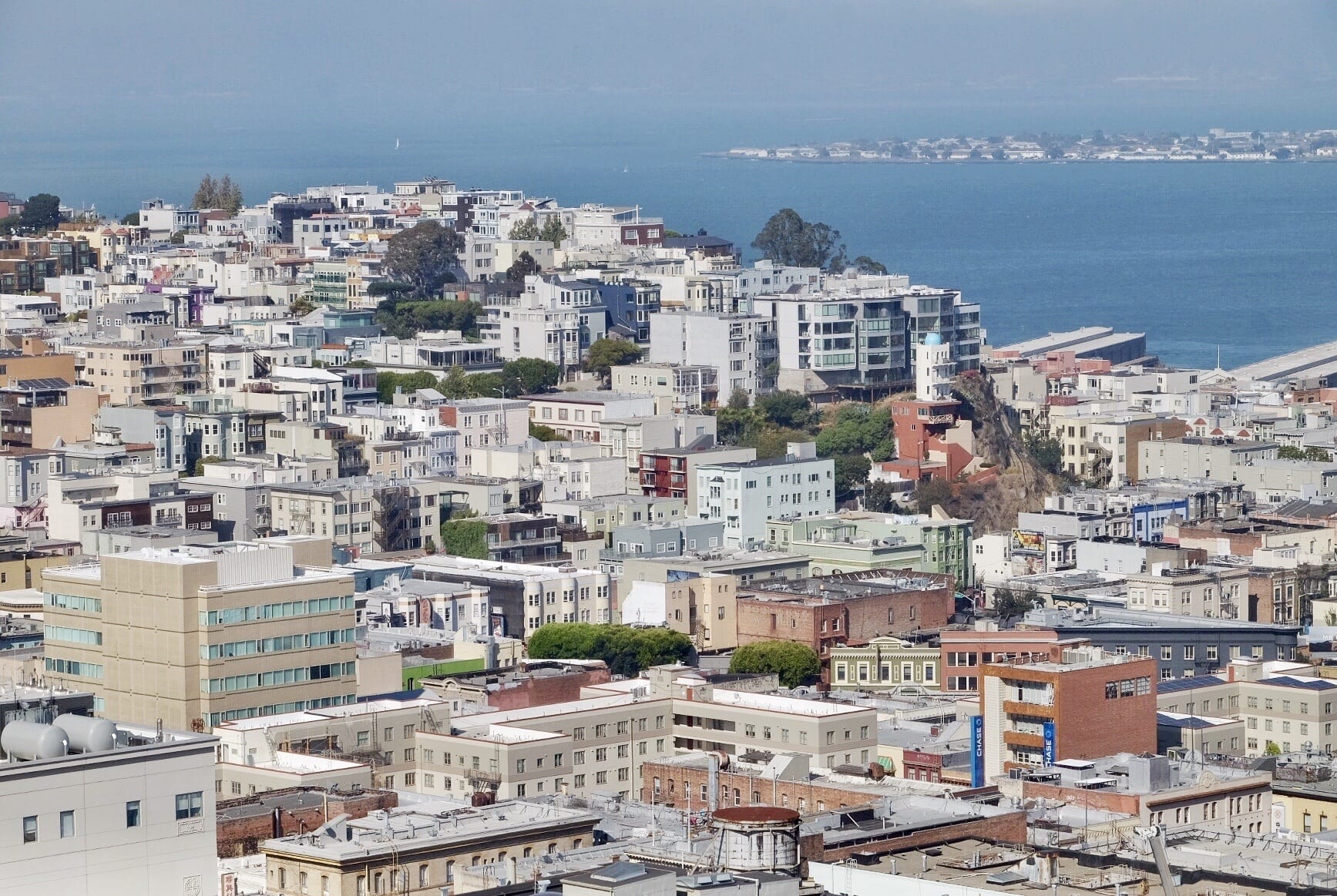 The San Francisco neighborhoods of Russian Hill and North Beach have square-shaped buildings stacked on top of each other, ascending and descending down the hills with occasional trees. In the background is San Francisco Bay and you can see Oakland through the haze in the distance.