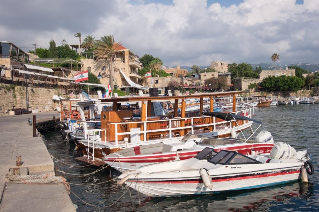 A row of small white boats docked at the edge of a stone pier, the sand-colored town of Byblos in the background.