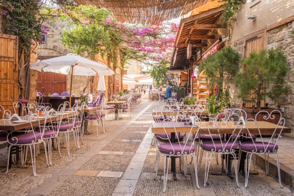 A courtyard in Byblos, Lebanon, with lots of wooden tables with white chairs. There are umbrellas and lots of plants. It feels like you're in the middle of a secret sandy room.