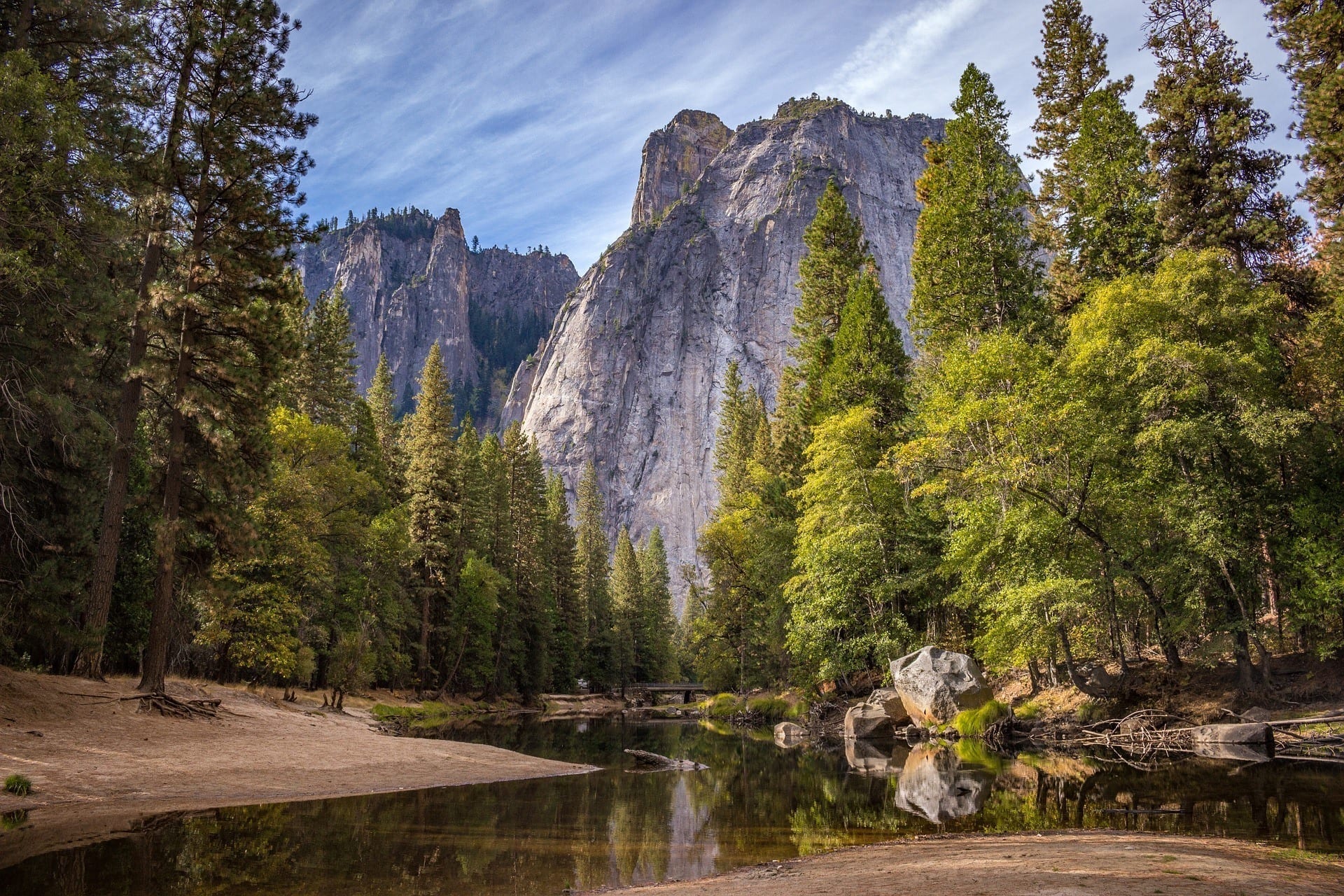 Yosemite National Park has huge looming mountains in the background underneath a blue and white streaky sky. There are bright green evergreen trees and water on the ground amongst the brown land.