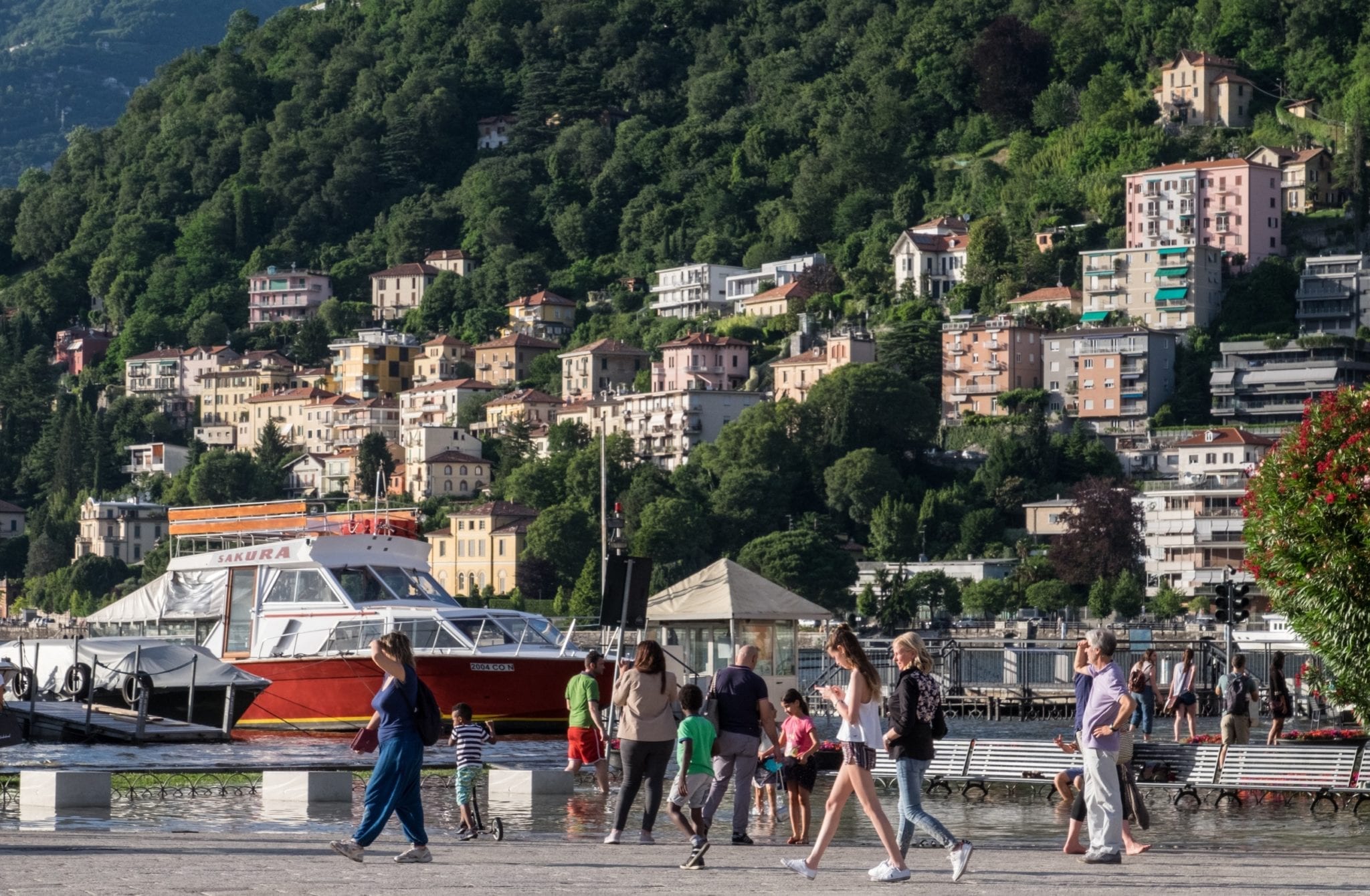 People walk on a piazza in front of buildings nestled into the hills of Lake Como.