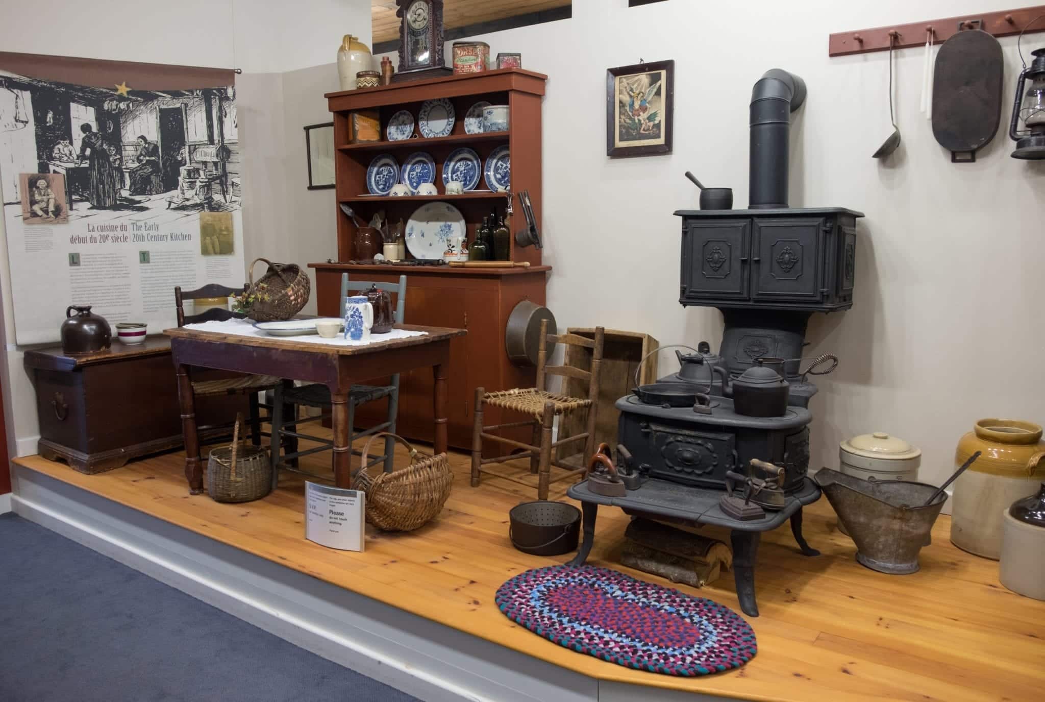 A recreation of an Acadian home at Les Trois Pignons with a china cabinet, wooden dining table, iron stove, and braided rug on the ground.