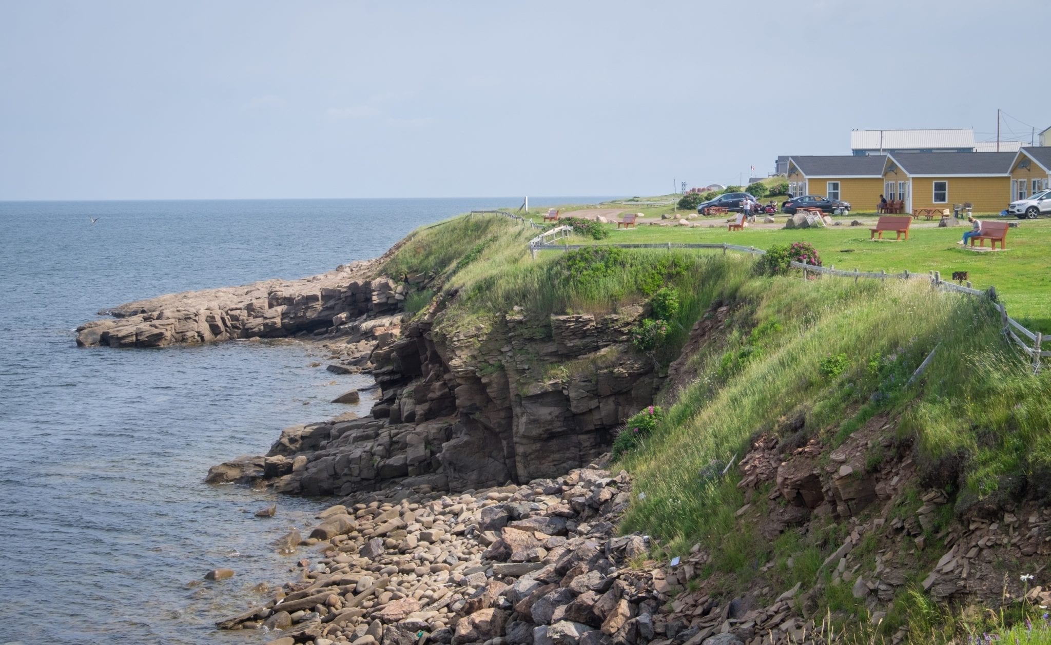 A rocky coastline falls into the sea. On the top is grass, two yellow cottages, benches, and picnic tables.