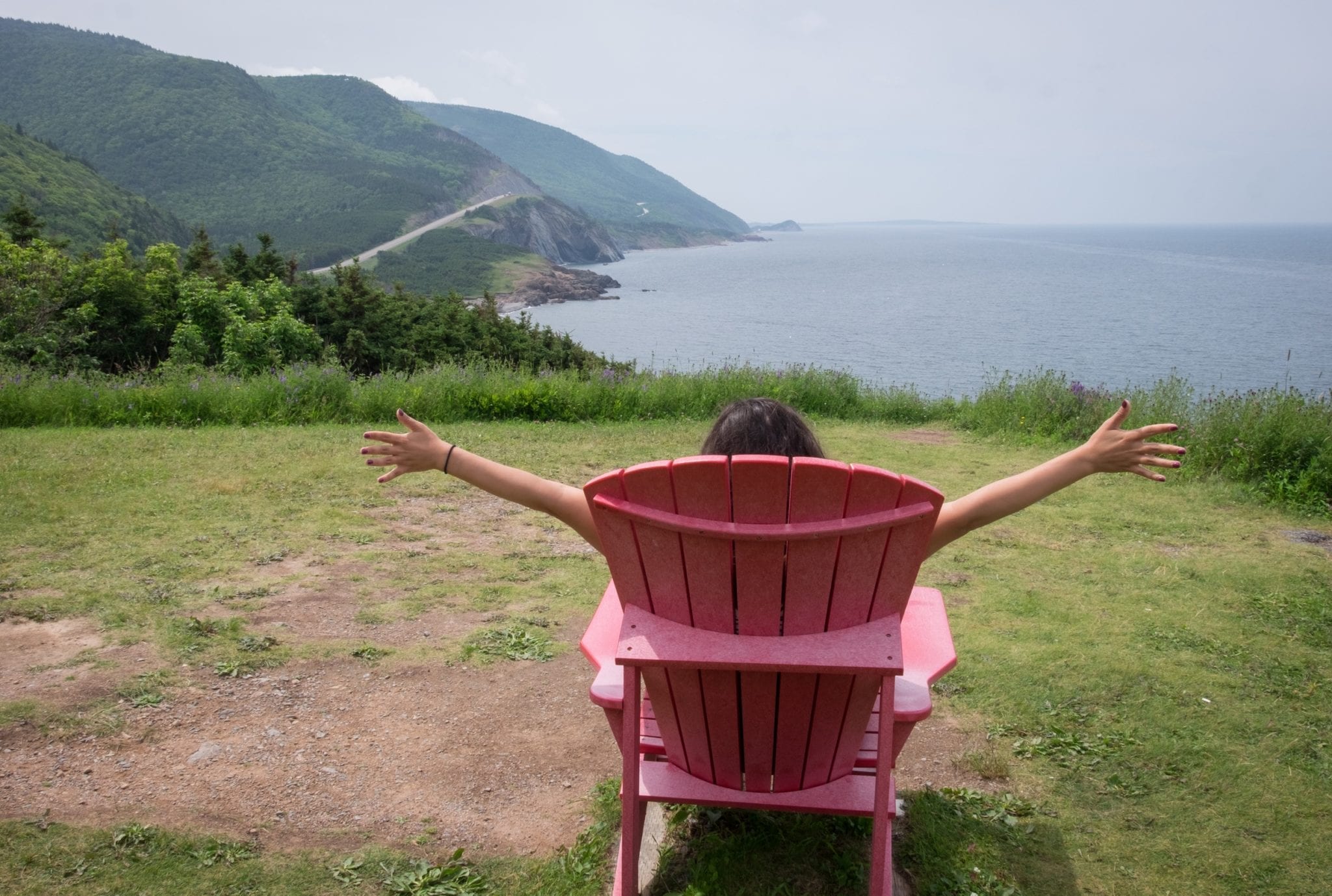 Kate sits in a red chair and has her arms up giving double thumbs ups. In front of her are hills, winding roads, and the ocean at Cape Breton Highlands National Park.