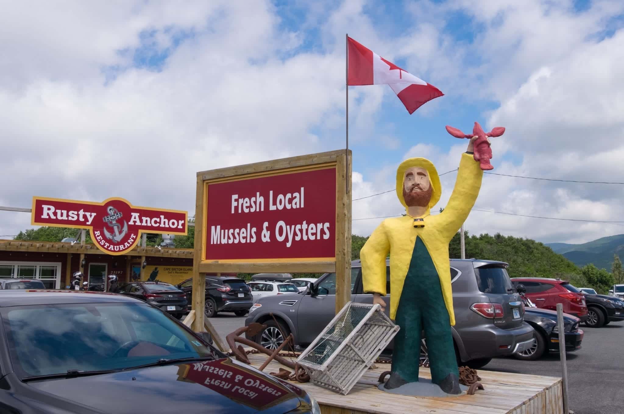 A wooden fisherman holds up lobster outside the Rusty Anchor restaurant in front of a sign that reads Fresh Local Mussels and Oysters.