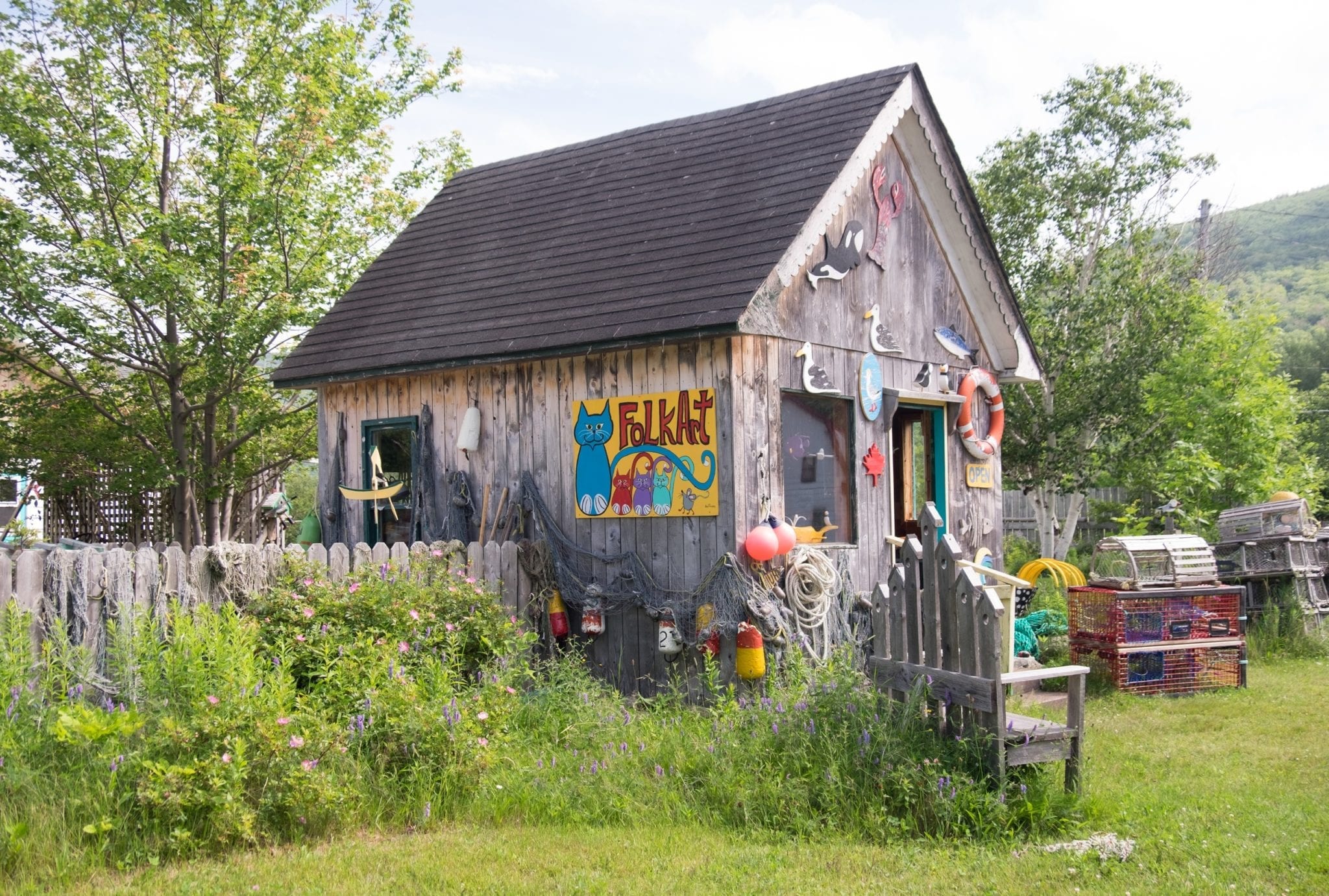 A small cabin covered with wooden art and brightly painted signs, surrounded by a fence overgrown with grass.