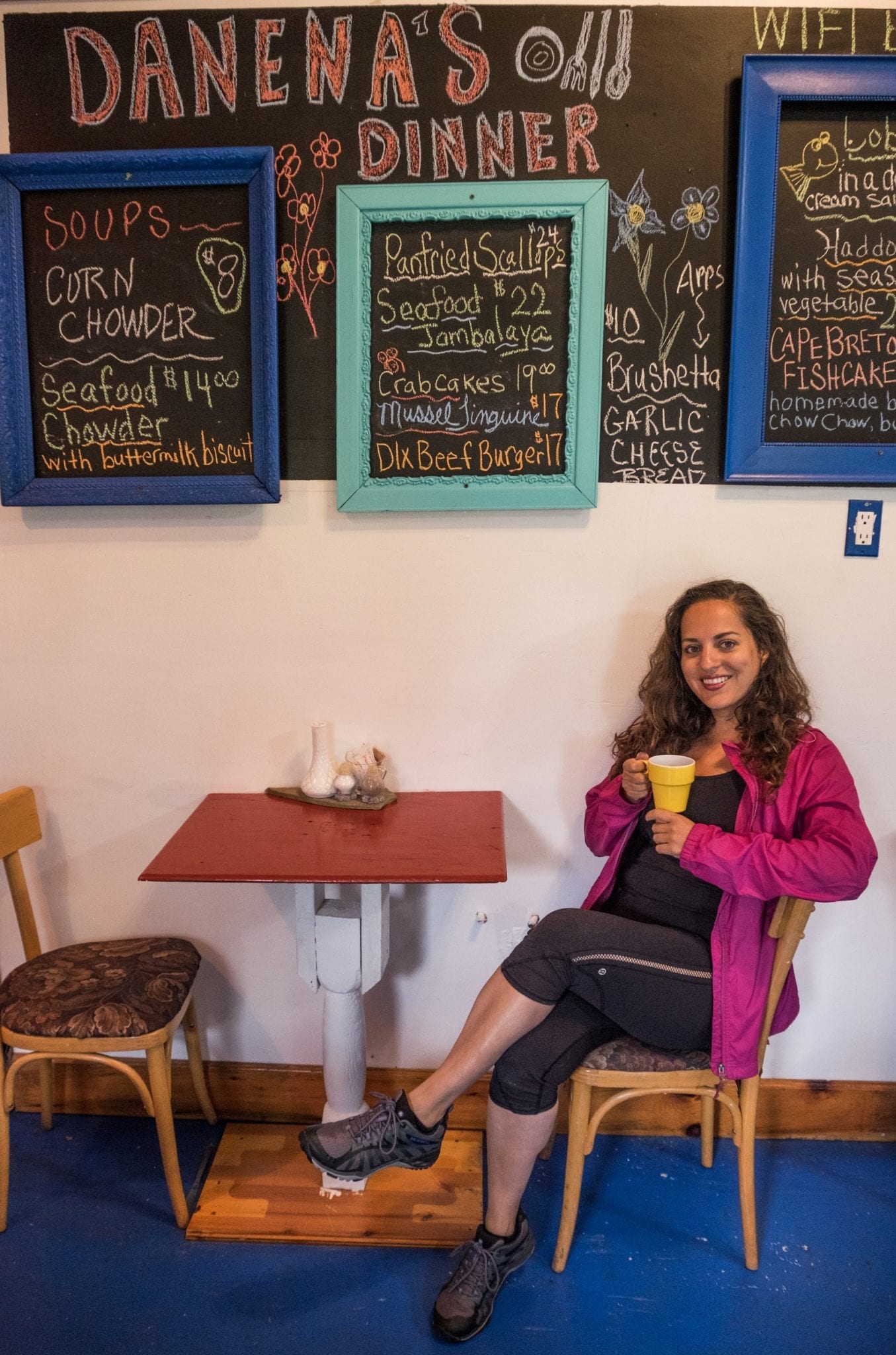 Kate wears a pink coat and smiles while holding a cup of coffee at a table beneath a blackboard detailing the restaurant's specials.
