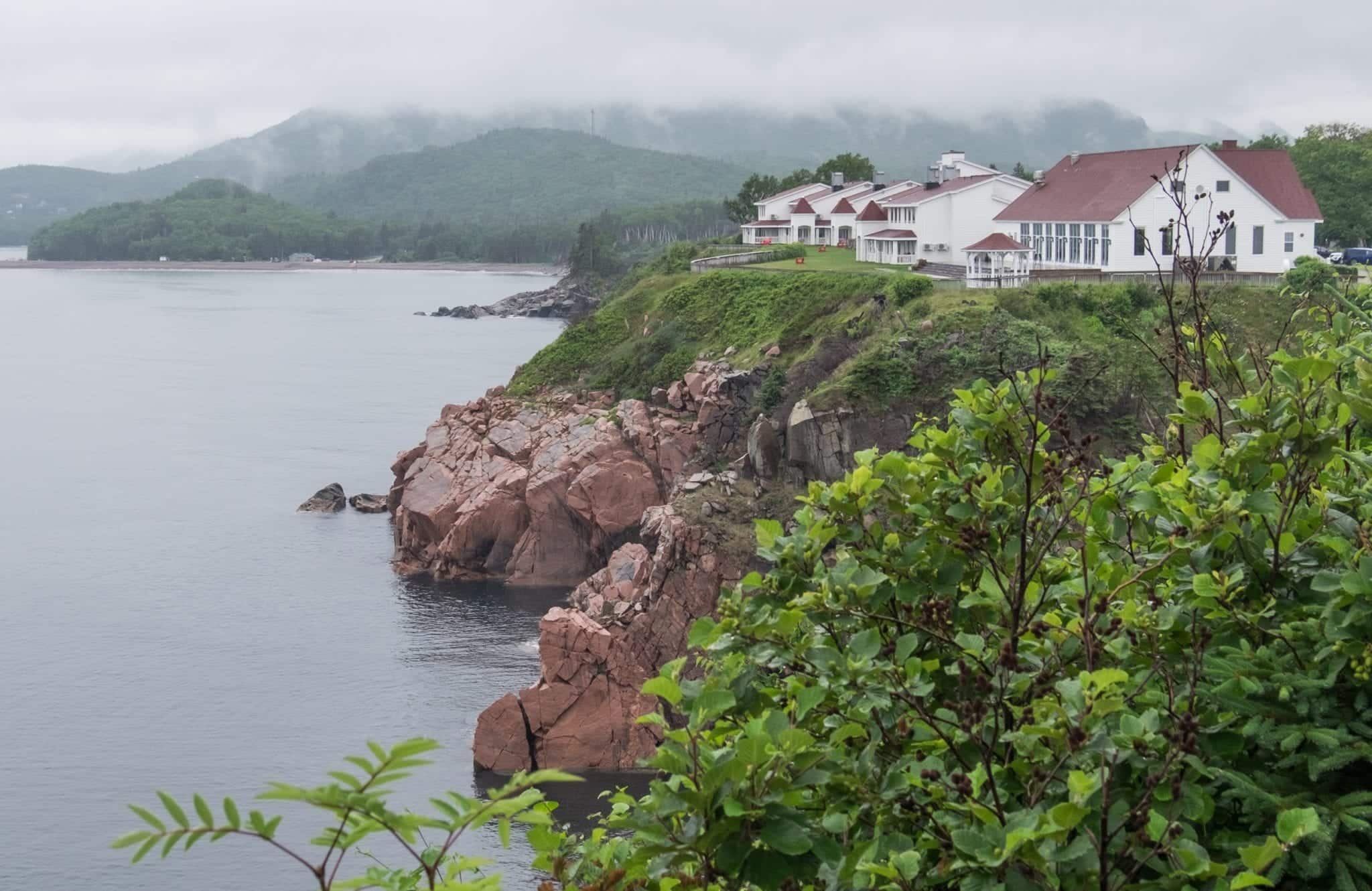 White cottages on top of red-brown cliffs among green vegetation, looking mysterious in the mist.