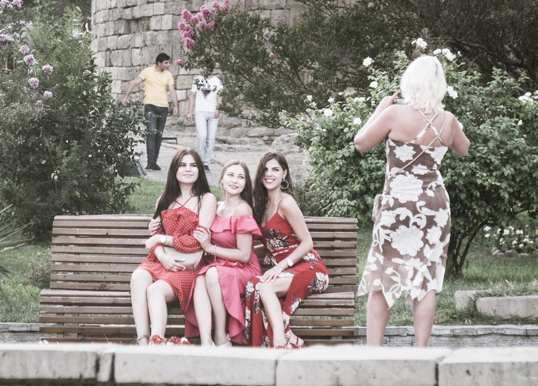 Three teenage girls in pink and red dresses sitting on a bench in the old city of Baku as a woman takes their photo.