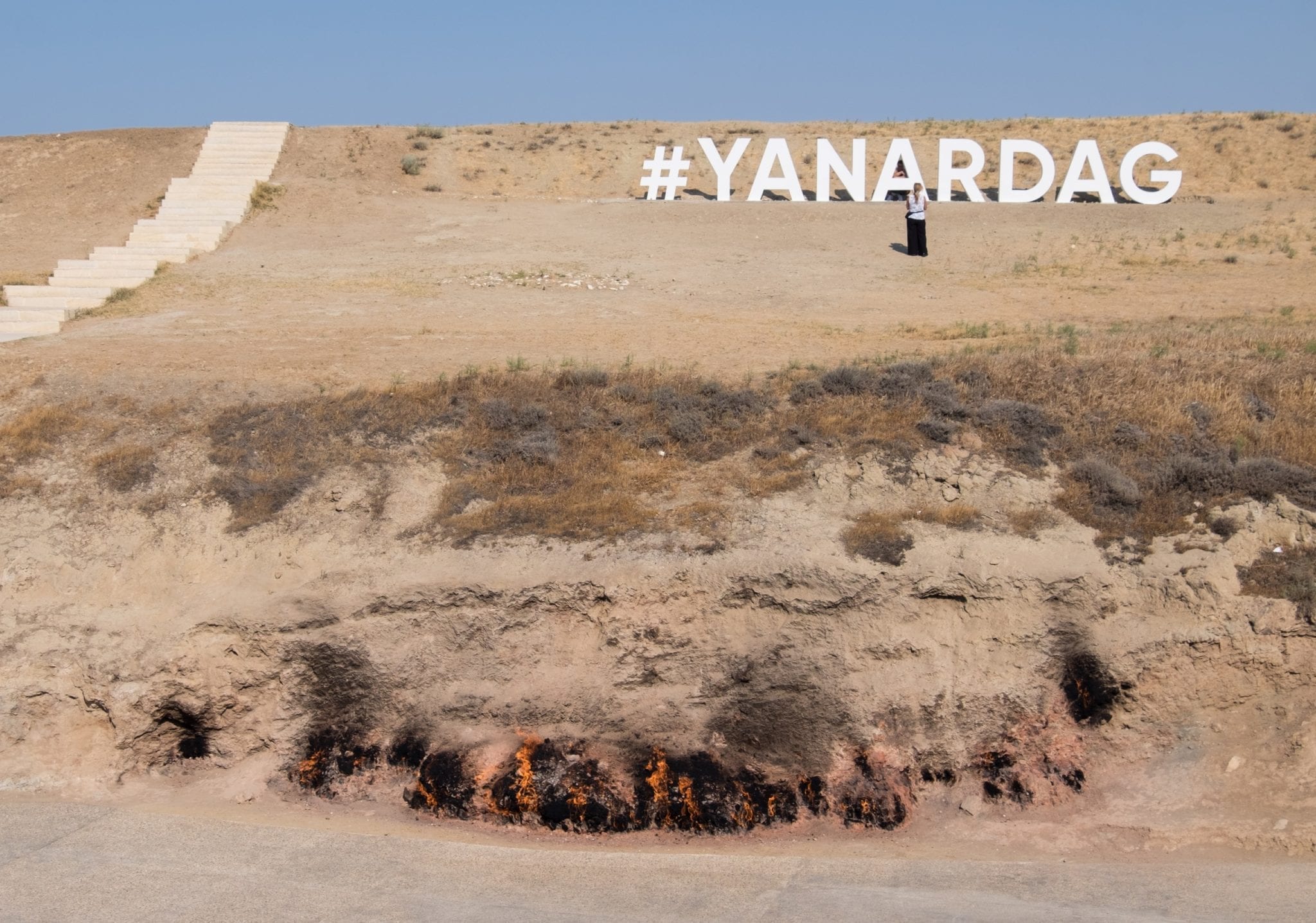 A brown hillside at Yanar Dag where flames burst out of the earth at the bottom. It says #YANARDAG in white on the hill, in the style of the Hollywood sign.