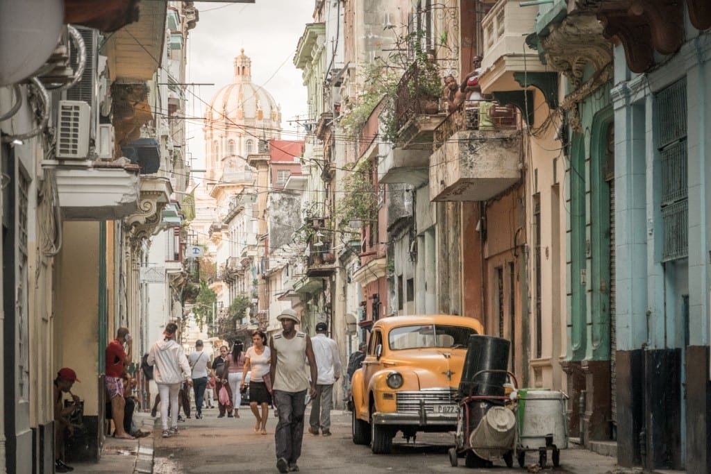 A street in Havana with people walking down it, lots of overhanging balconies, and a bright yellow classic car.
