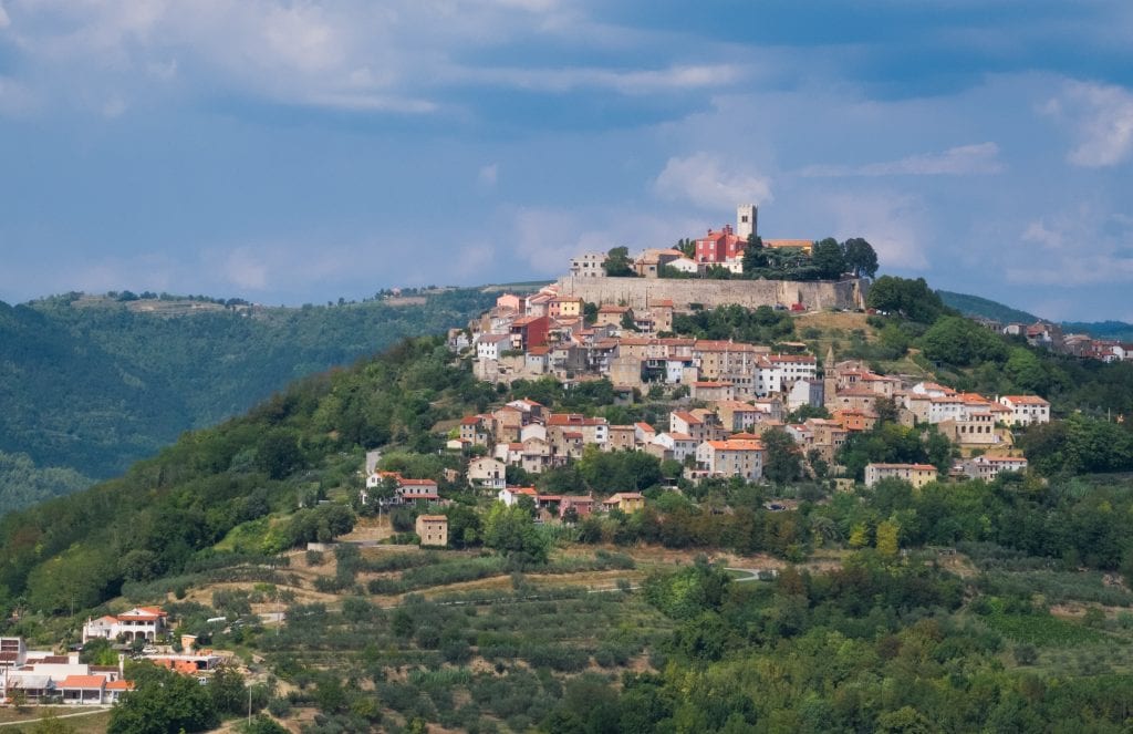 The mountaintop town of Motovun, warm-colored stone buildings perched on top of a hill, underneath a blue and white sky.