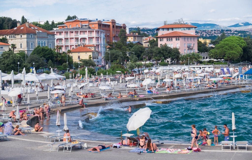 Beachgoers sitting on a concrete slab on the edge of a bright blue ocean in Opatija, Croatia, with Hapsburg-style ornate pink and orange buildings in the background.
