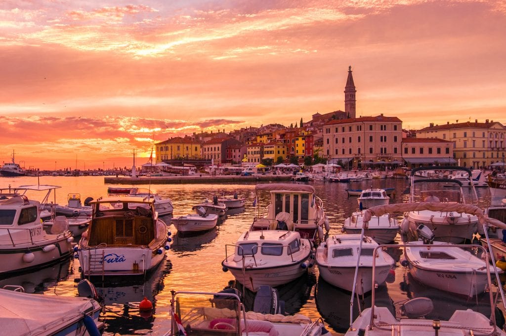 Rovinj at sunset: The whole sky is lit up orange and yellow. In the foreground are rows of small white boats docked up; in the back is the old city of Rovinj on the edge of the water, a big church tower sticking out of the top.