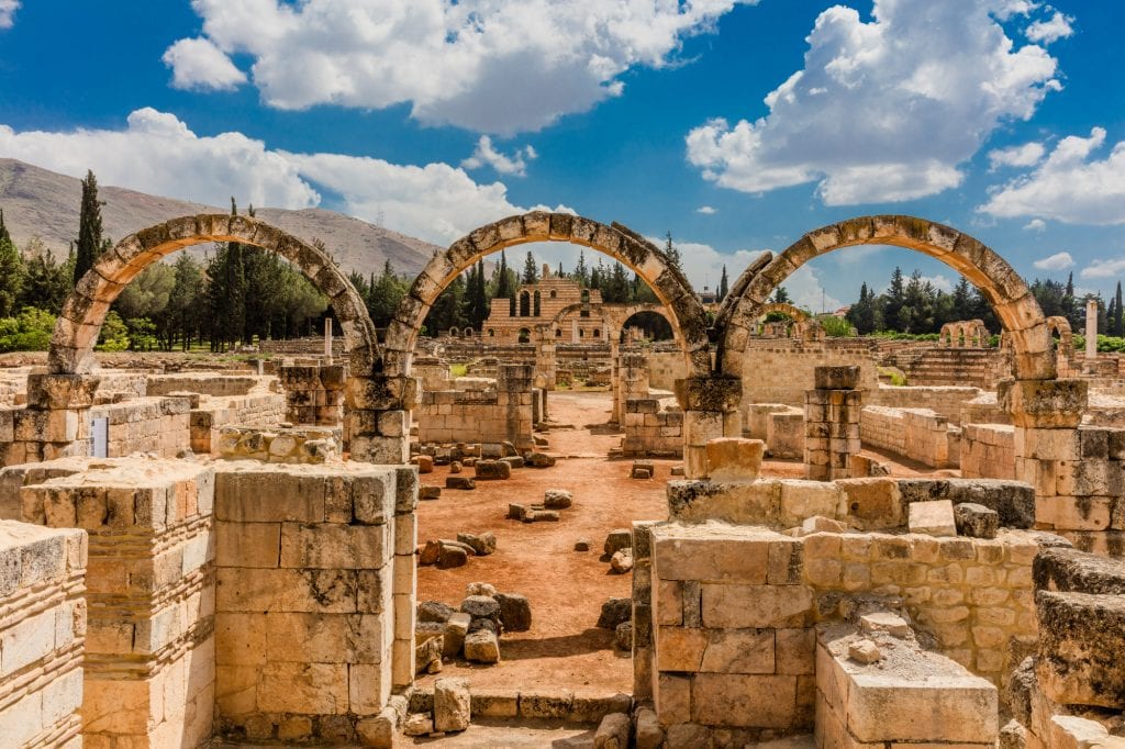 The ruins of Anjar: you see broken-down stone buildings. In the middle are three stone arches, each made from a single row of stones. In the background, mountains (and beyond them, Syria).