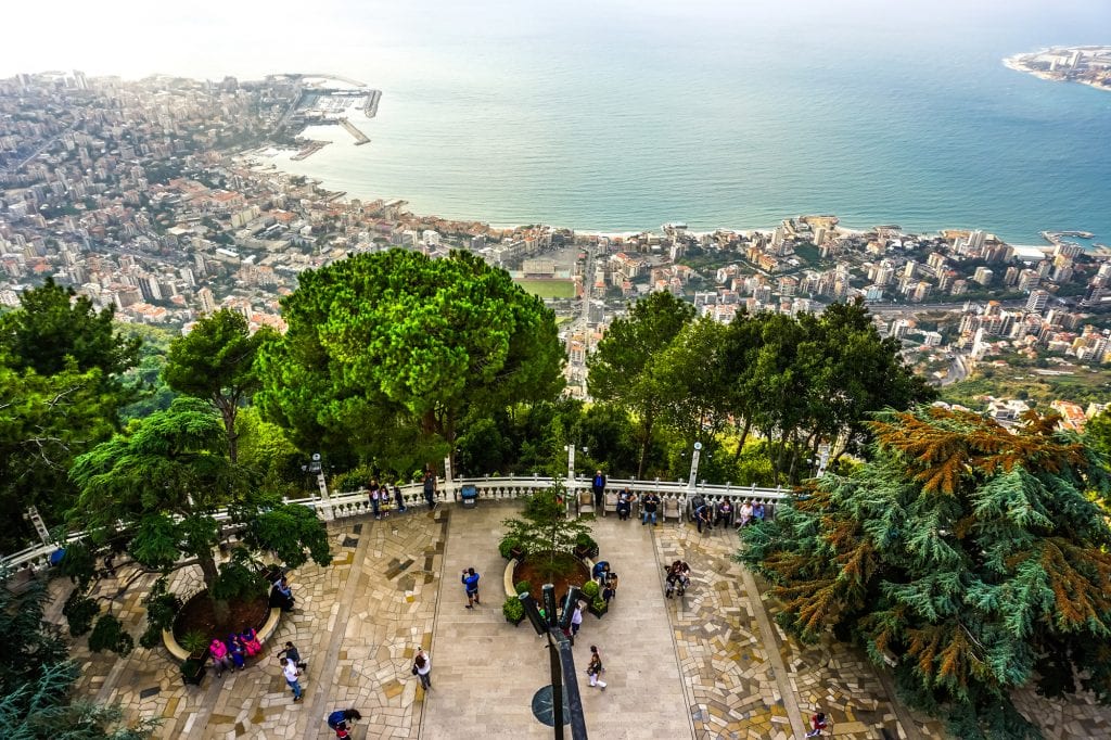 In the bottom of the photo, up close, you see people taking photos on a stone platform. Beyond them, a row of trees, then you see way below them in the distance is the city of Jounieh along the turquoise water.