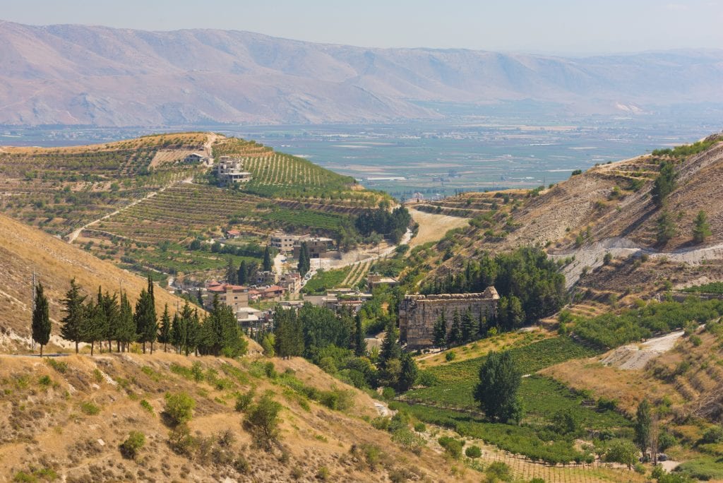 A mountainous landscape in Lebanon. On the small hills are terraced vineyards and small buildings; in the distance, you see a steep mountain.