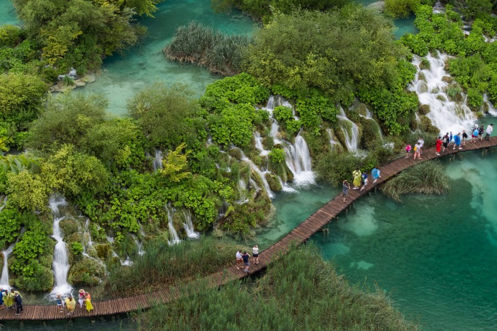 An aerial shot of the Plitvice Lakes. A wooden platform with people walking on it sneaks through the clear teal lakes. Next to the platform are something like 20 mini waterfalls, all bursting through the trees and bushes.