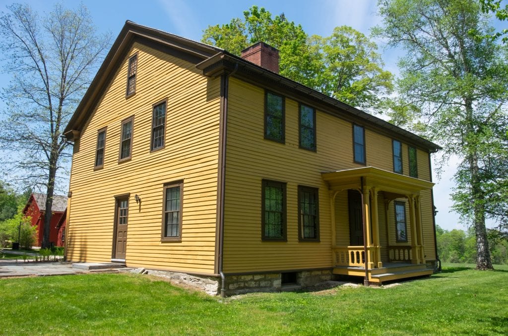 A mustard yellow house with brown trim and a small porch in front, set among trees.
