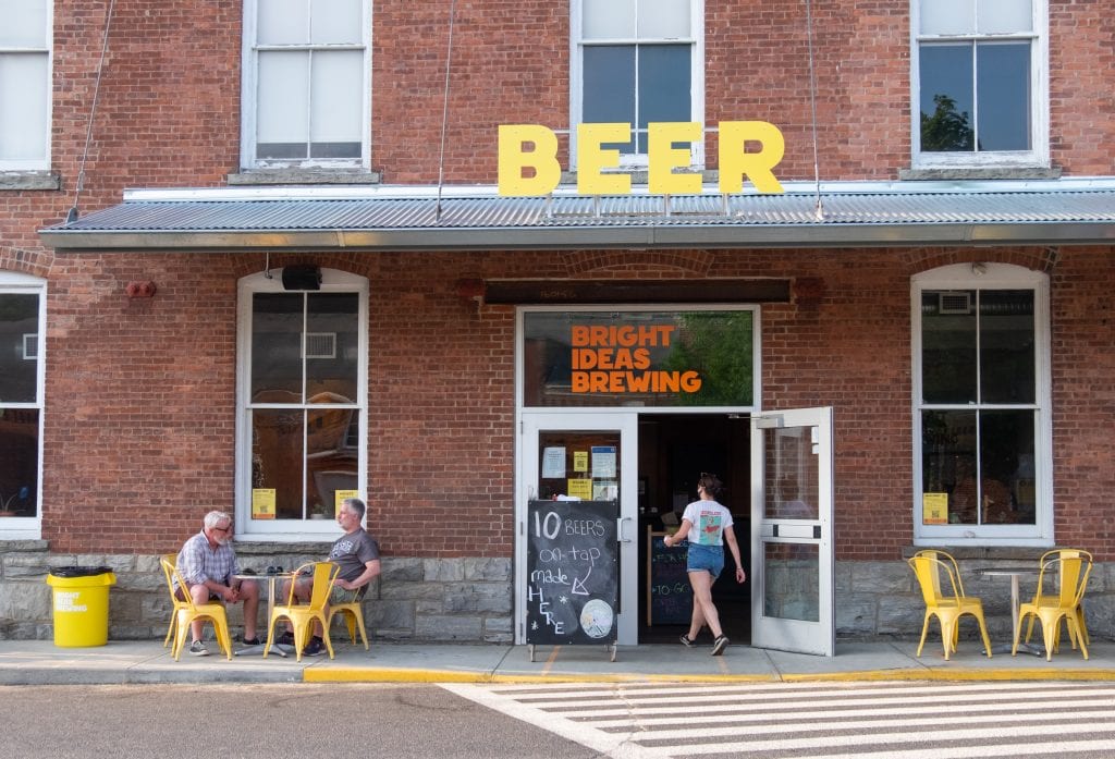 A brewery in a brick building. A big yellow sign reads BEER on top; you see two older man sitting at two bright yellow chairs outside, as a waitress walks into the front door.