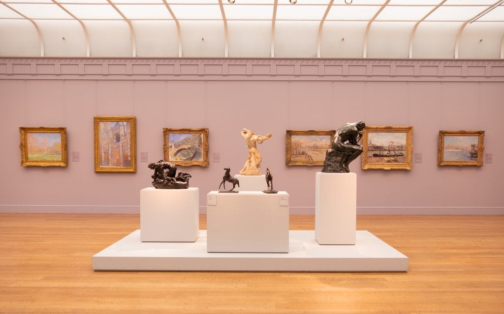 A pale lavender room at the Clark Institute with several gold-framed Impressionist paintings on the walls: in the foreground, several marble and metal sculptures. Two are of small horses; one is a man sitting and thinking with his head on his hand.