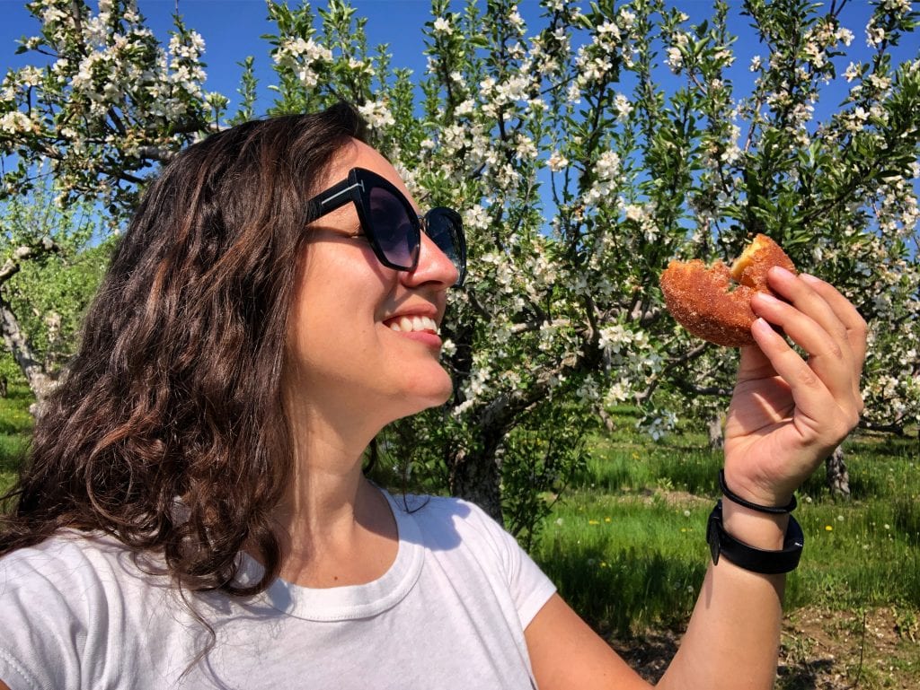Kate holds a half-eaten cider donut in her hand and grins at it lovingly. Behind her are blossoming apple trees.