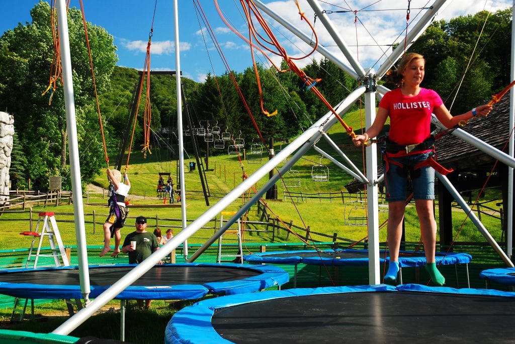 Two kids jumping on trampolines while wearing harnesses at Jiminy Peak Mountain Resort. In the background, empty ski lifts.