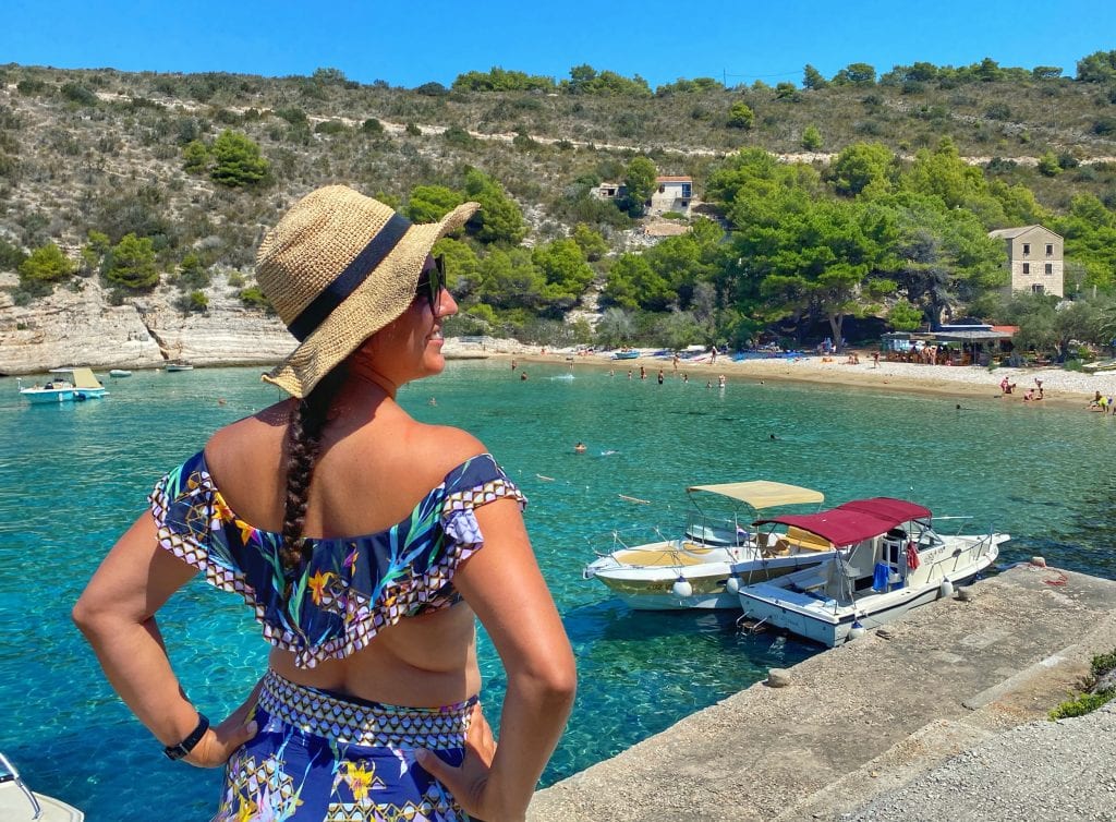 Kate standing in front of the bright blue-green water of the Adriatic. She faces away from the camera and wears a straw hat and sunglasses, a navy ruffled bikini top with tropical flowers on it, and harem pants that match the top.