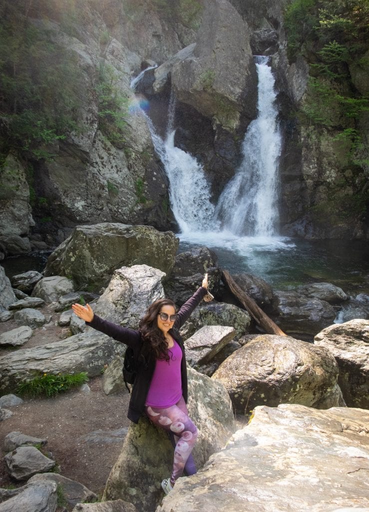 Kate standing in front of the two-waterfall Bash Bish Balls in Massachusetts, wearing a purple shirt and leggings and her arms up in the air.