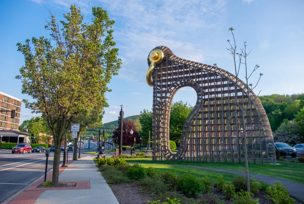 A modern sculpture that looks a bit like a bird built from an iron grid, a golden handle serving as beak and eyeball, overlooking a busy street in North Adams.