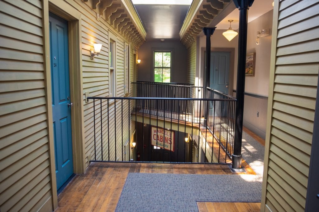In the hallway, you see several entrances to rooms. It's an open concept and you can see the floor below, which has a vintage sign reading "Marcel's Cabins - Color TV."