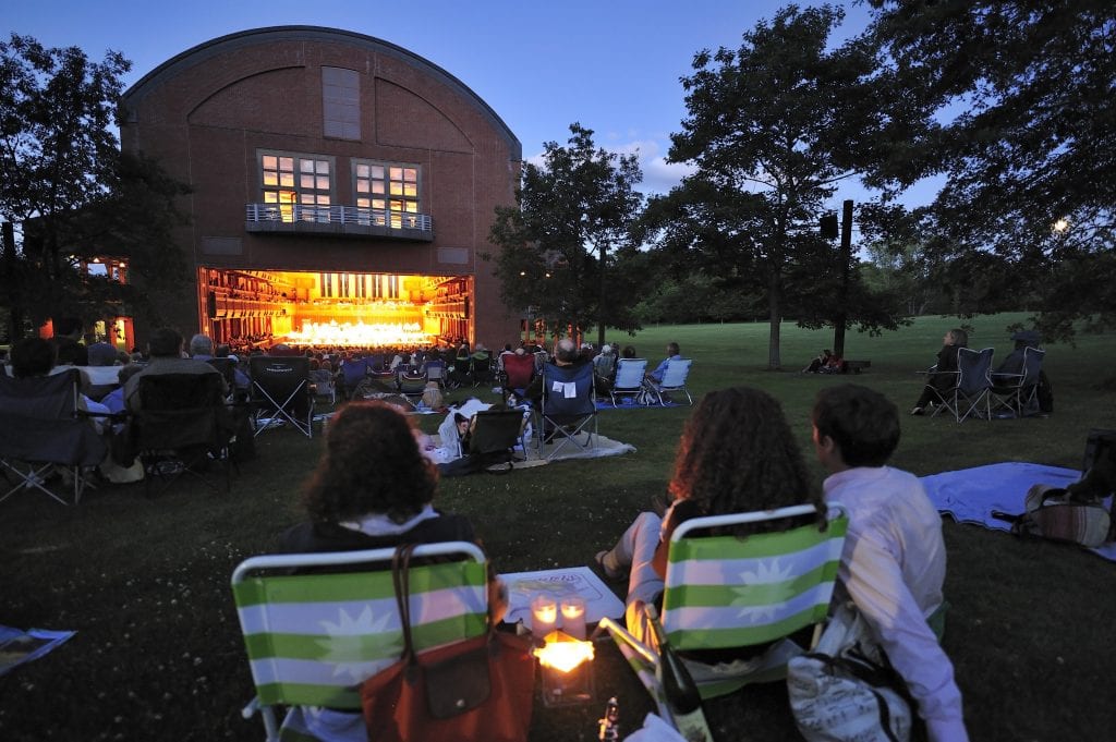 The red brick theater of Tanglewood with a big open door; in front of it are people sitting in lawn chairs, listening to the music on a summer night.