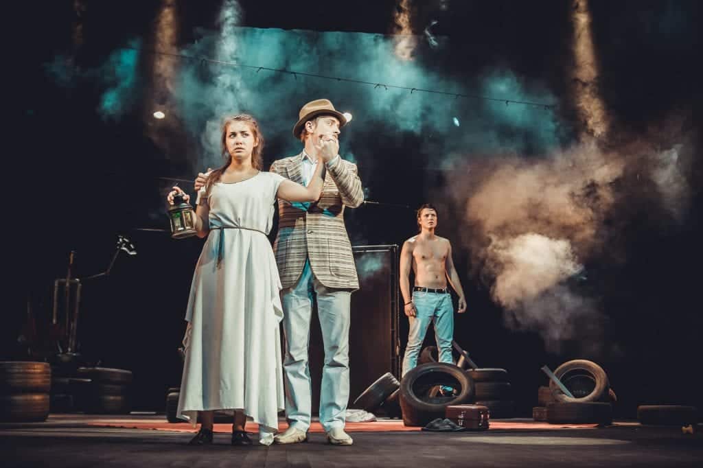 A scene from a play, a man in a hat and blazer holding the hand of a woman in a nightgown holding a lantern. For some reason there's a shirtless dude in the background and I'm not complaining.