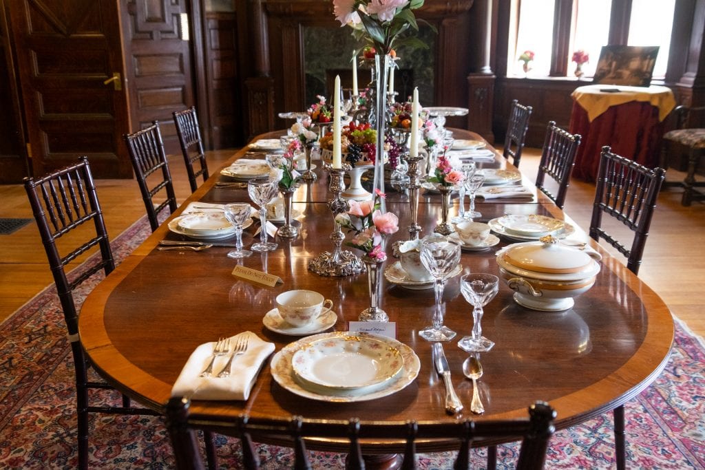 A fully set dining table at Ventford hall covered with gold-tipped china, crystal glassware, silver candlesticks, and silver vases filled with pink fake flowers.