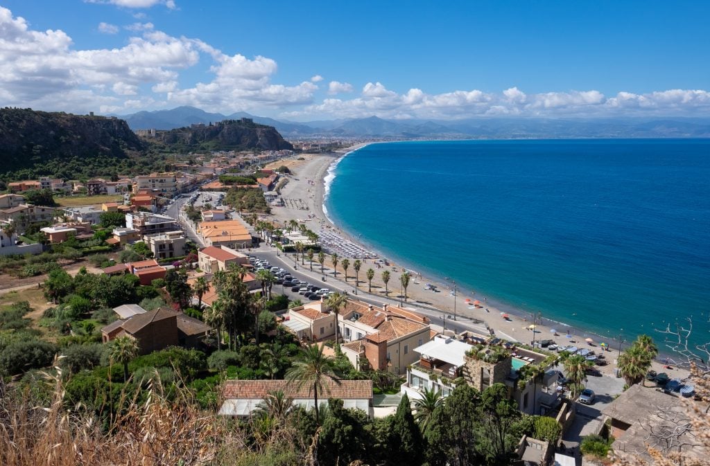 Milazzo: on the right, a half-moon of calm gray beach next to bright blue water. On land, houses and vegetation, and in the background, you see what look like small mountains (the ridge), one topped with the ruins of a castle.