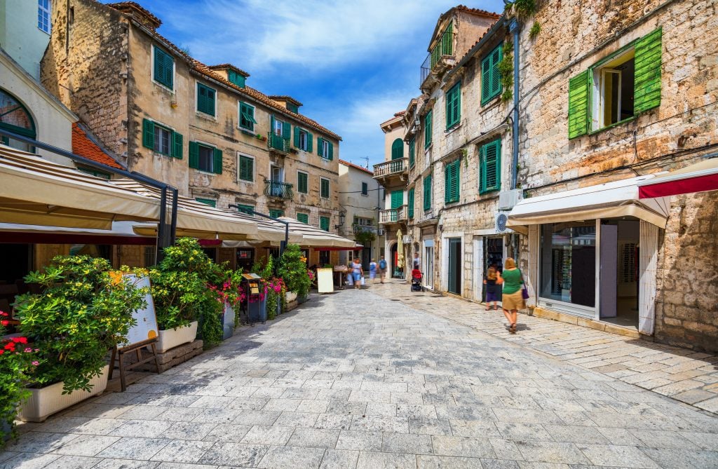 A street in Split's old town with stone buildings with emerald green shutters, and cafes surrounded with plants for privacy.