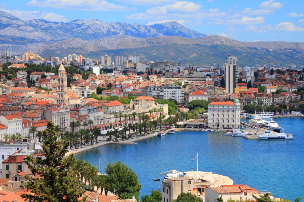 An aerial view of Split, a city with lots of white buildings, orange roofs, and a big church bell tower pushed up against a palm tree-lined shore, mountains in the distance behind.