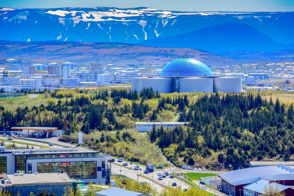 Perlan: a building made of tall gray cylinders with a glass dome on top. It's surrounded by pine trees and in the background are lots of apartment buildings and a view of mountains.