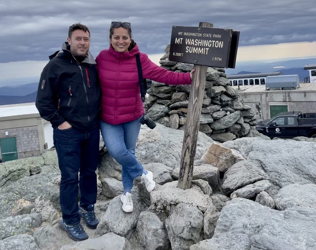 Kate and Charlie standing side by side on a rocky top with a sign reading Mount Washington Summit.